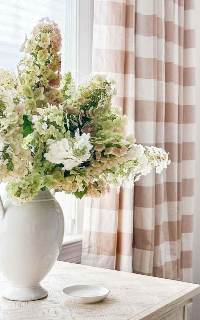 beautiful bouquet of drying pink and green hydrangeas in a white urn on a table with light brown and white buffalo check curtains