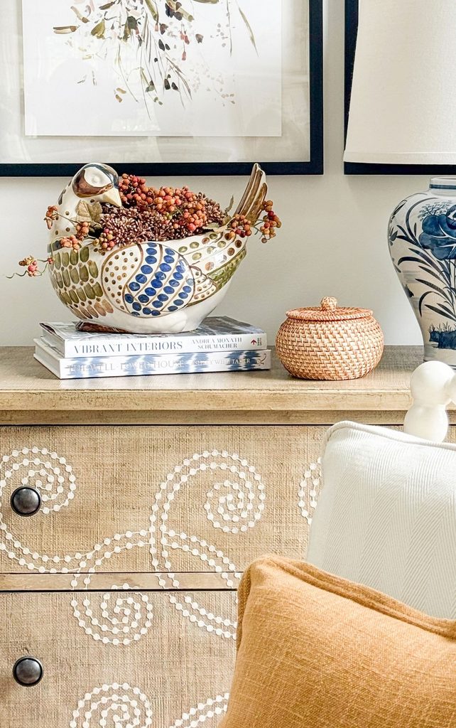 Hand-painted ceramic pheasant bowl filled with fall berries on a stack of books beside a blue and white lamp, rattan box, and pillows
