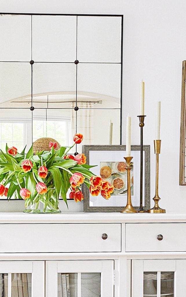 Brass and bronze taper candlesticks in varied heights on a white sideboard with orange tulips, a botanical print, and a grid mirror