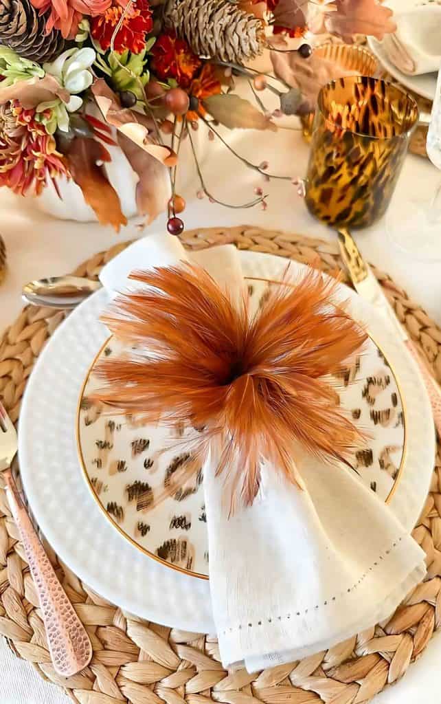 Thanksgiving place setting with a woven charger, white and animal print plates, a white napkin with a rust feather napkin ring, a tortoiseshell goblet, copper flatware, and a fall floral centerpiece with pinecones
