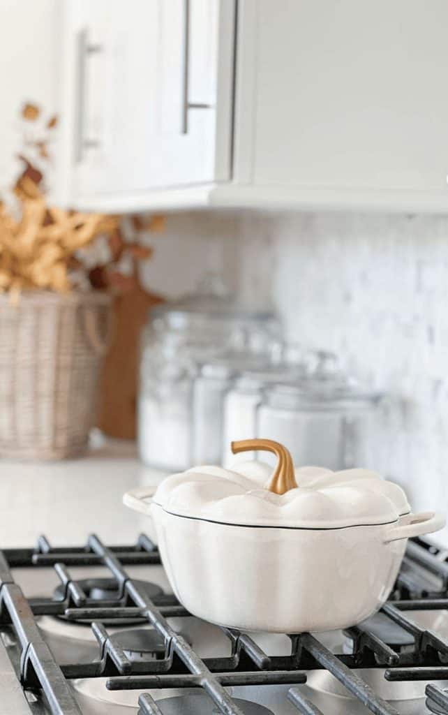 Creamy white pumpkin Dutch oven with a gold stem lid on a gas stove, with glass canisters, a marble backsplash, and a basket of fall stems in the background