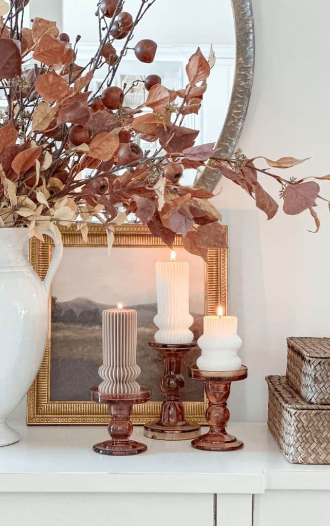 Three amber glass candlesticks holding lit ribbed pillar candles in front of a gold framed landscape painting, with fall stems in a white pitcher, a round mirror, and woven boxes on a white mantel