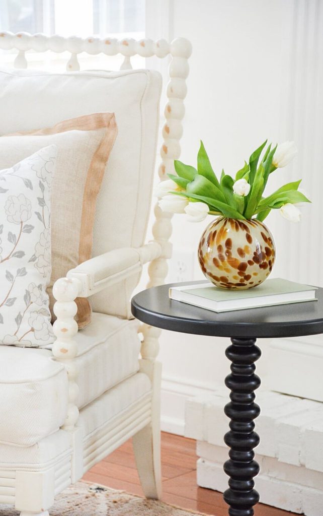 Small black spindle drink table styled with a book and an amber glass vase of white tulips beside a white spool chair