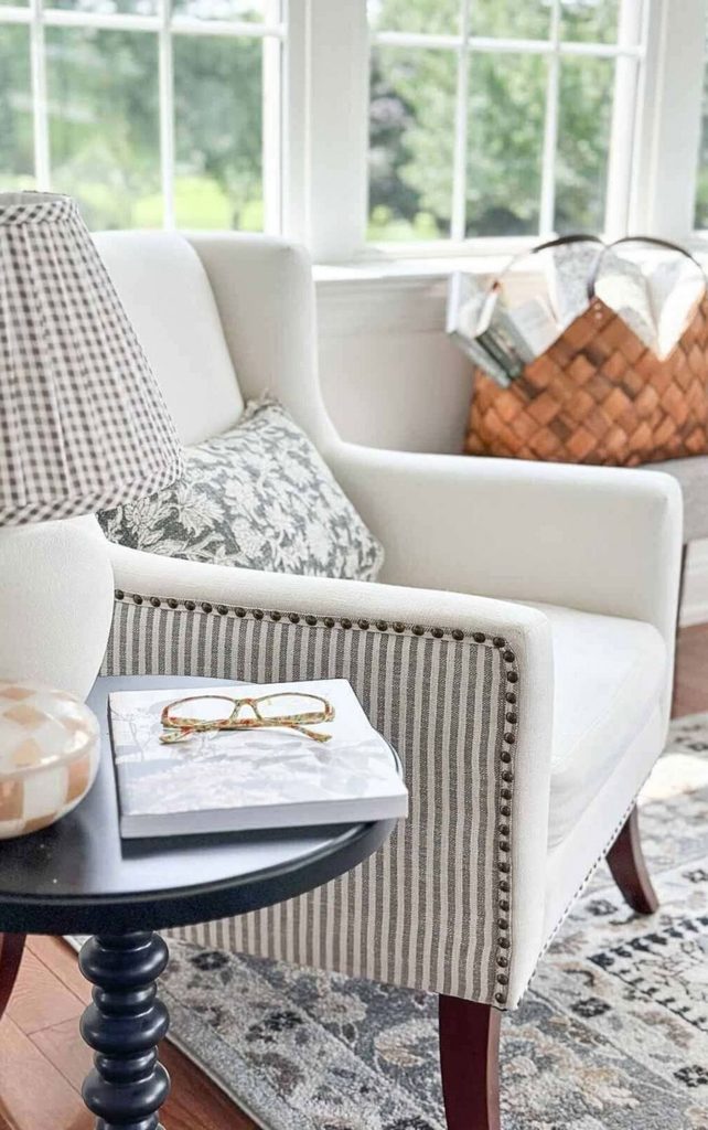 Small round black spindle side table holding a gingham shaded lamp, a book, and reading glasses beside a white chair with a striped arm and nailhead trim