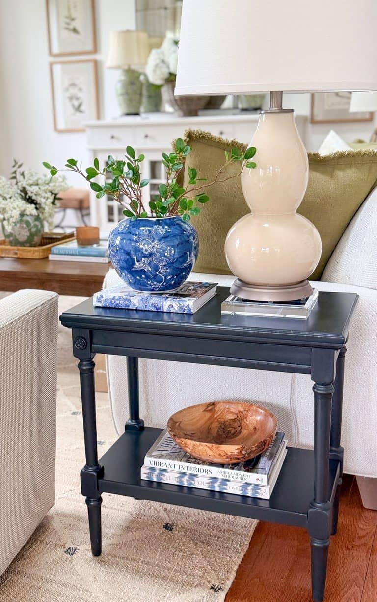 Deep blue end table beside a white sofa styled with a cream gourd lamp on an acrylic base, a blue and white vase of greenery on a book, and a wooden bowl on stacked books on the lower shelf