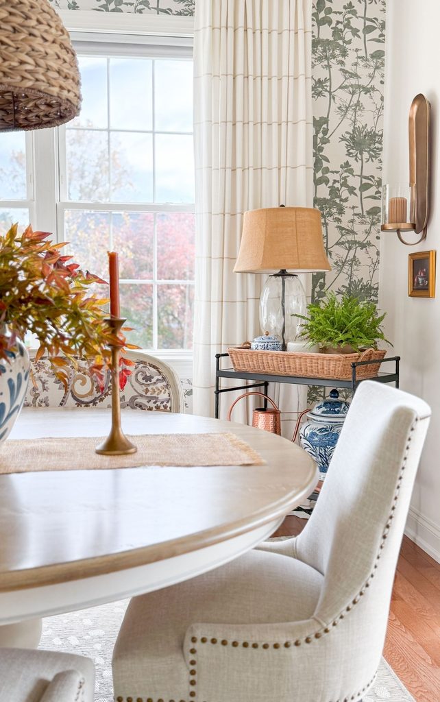 Dining room corner with green botanical wallpaper, a brass candle sconce, and a table lamp on an iron cart with a fern and blue and white ginger jars, giving soft, warm Sunday Home lighting