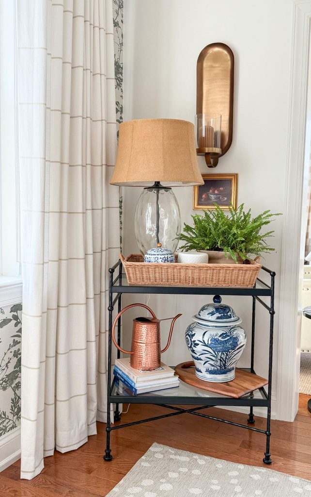 Iron and glass cart styled with a table lamp, a fern in a wicker tray, a copper watering can, stacked books, and a blue and white ginger jar beside botanical wallpaper in a Sunday Home dining room