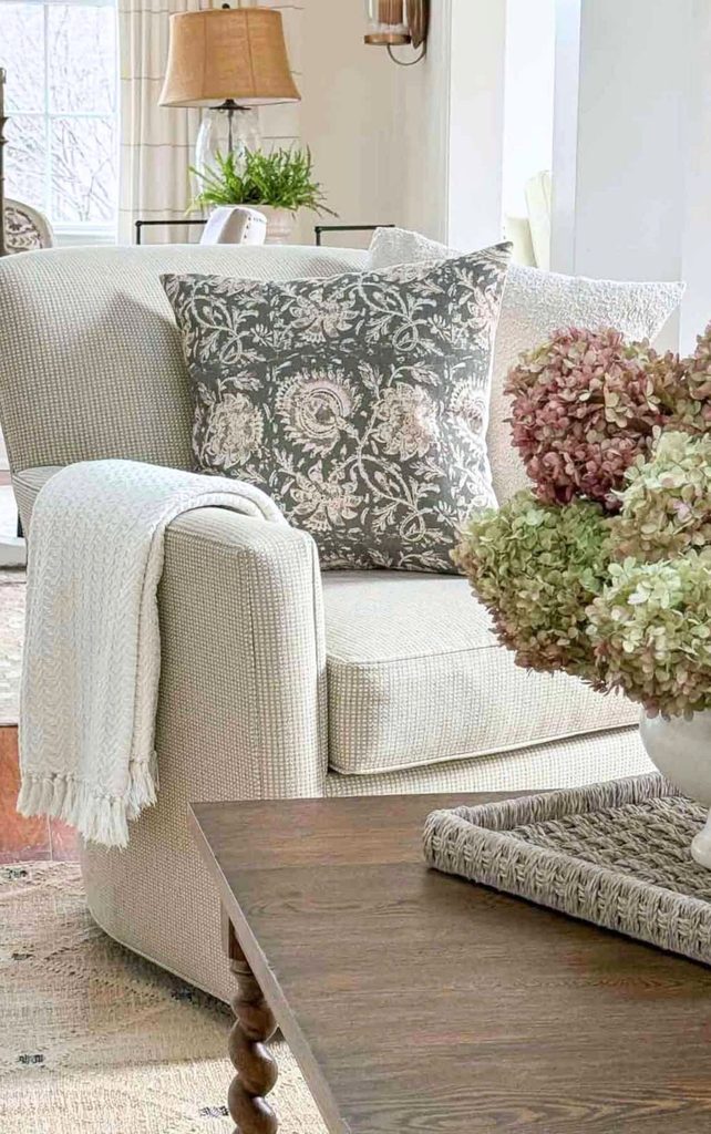 Upholstered armchair layered with a cream fringed throw, a gray floral pillow, and a textured white pillow beside dried hydrangeas on a wood coffee table, showing cozy Sunday Home textiles