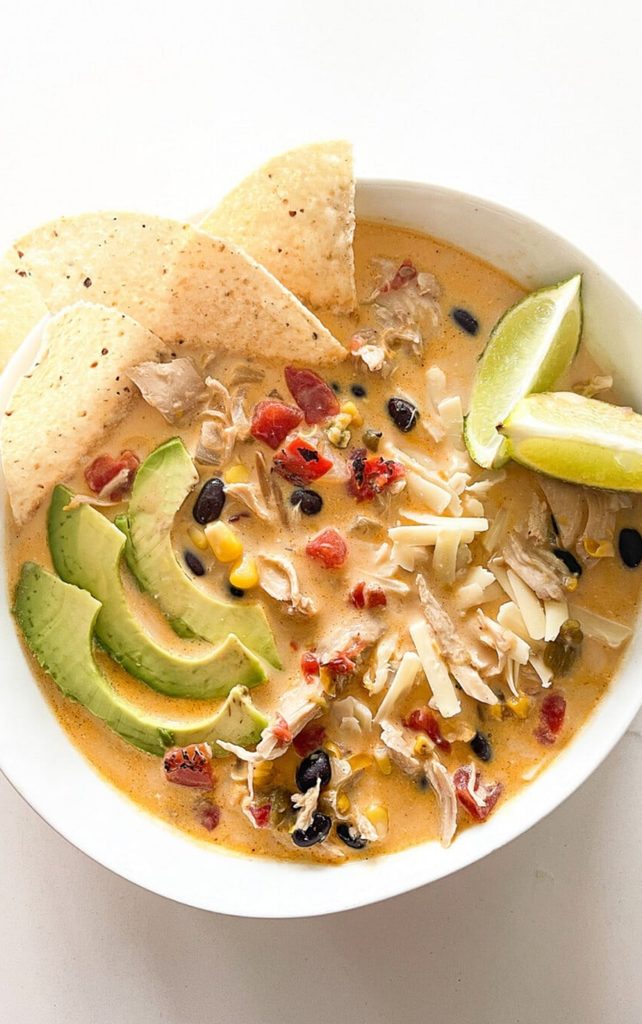 Bowl of creamy chicken soup with black beans, corn, and tomatoes, topped with sliced avocado, shredded cheese, lime wedges, and tortilla chips, a simple Sunday Home meal