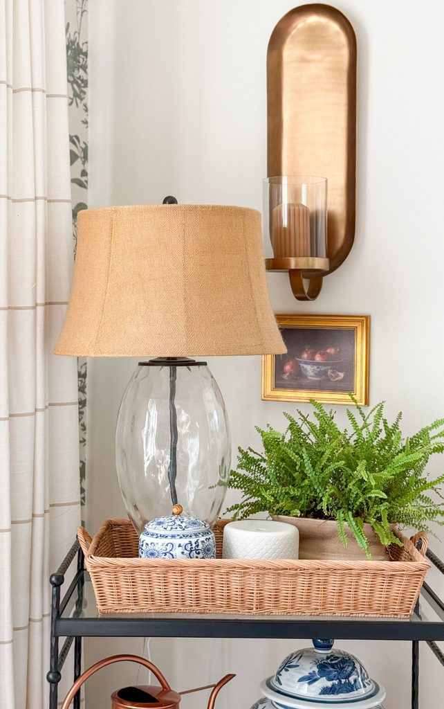 Brass candle sconce above a glass table lamp with a burlap shade, a Boston fern, and blue and white jars in a wicker tray, showing soft, warm Sunday Home lighting
