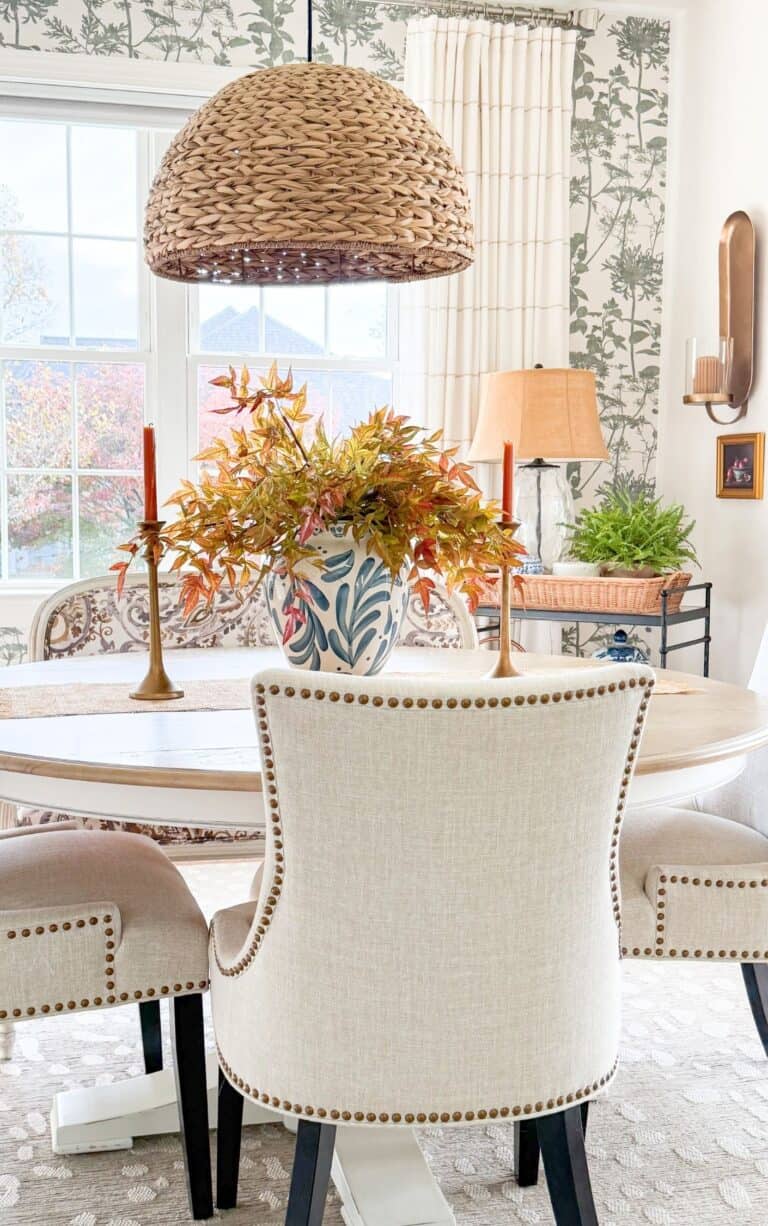 Round breakfast table by a kitchen banquette with fall branches in a blue and white vase, brass candlesticks, and a warm wall sconce, showing the Sunday Home trend