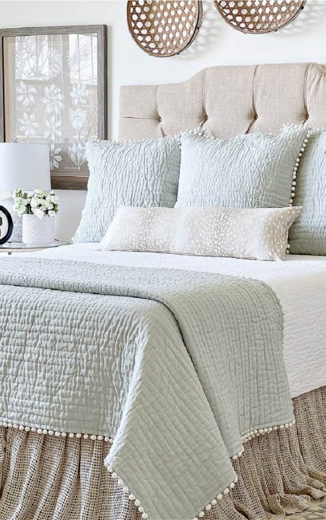 Neatly made bed with a tufted linen headboard, a sage quilt with pom pom trim, fluffed pillows, and white flowers on the nightstand, showing a bedroom after a simple Sunday reset