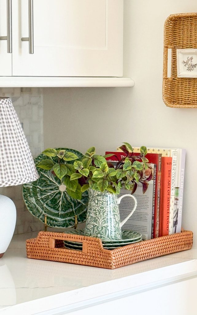 Kitchen counter with a rattan tray holding cookbooks, a green cabbage plate, and a green floral pitcher of fresh greenery beside a gingham lamp, showing a tidy Sunday reset
