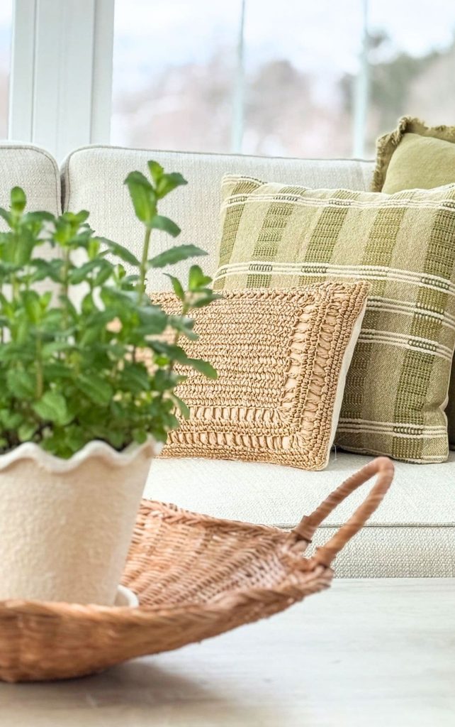 Cream sofa with fluffed green striped, olive, and woven pillows behind a potted mint plant in a scalloped pot and a wicker basket, showing a living room after a simple Sunday reset