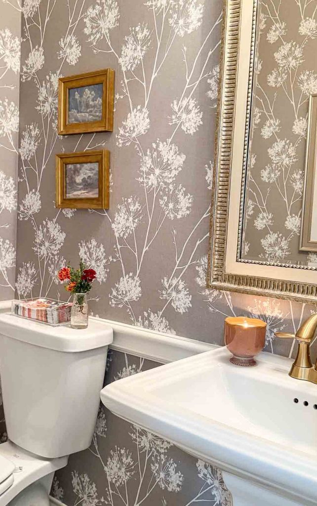 Powder room with gray and white botanical wallpaper, gold framed art, a lit candle by a pedestal sink, and a small jar of red and orange flowers with guest towels, showing a fresh Sunday reset