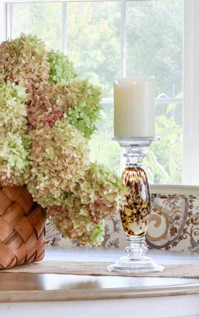 Glass candlestick with a tortoiseshell center holding a white pillar candle beside dried limelight hydrangeas in a woven basket